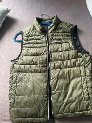 Olivgrön dunväst från Jack & Jones - Snygg olivgrön dunväst från Jack & Jones med quiltad design och hög krage. Västen har dragkedja framtill och två stora fickor på sidorna. Perfekt att slänga över en hoodie för extra värme. Materialet är lätt och smidigt.