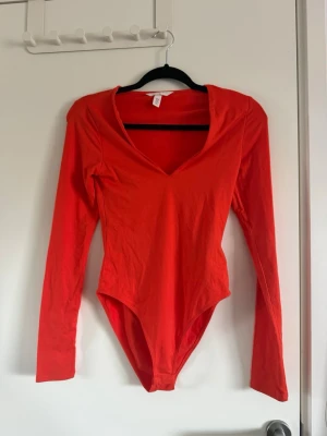 Orange långärmad body med v-ringning - Snygg orange body med långa ärmar och djup v-ringning. Tillverkad i mjukt stretchigt material som sitter skönt mot kroppen. Perfekt att styla med jeans eller kjol för en trendig look.
