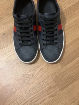 Gucci sneakers med röd och blå rand - Snygga svarta Gucci sneakers med diskret GG-mönster, röda och blå sidoremsor samt vit sula. Skorna har svarta skosnören och metallic-detaljer i rött och lila på hälen. Perfekta för dig som vill ha en klassisk men ändå unik look.