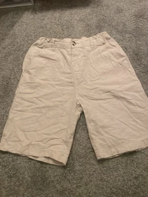 Beiga shorts från Kappahl - Säljer ett par beiga shorts från Kappahl med elastisk midja och knappstängning. Shortsen har bälteshällor och sidofickor, perfekta för sommardagar. Materialet känns mjukt och luftigt, vilket gör dem riktigt sköna att bära.