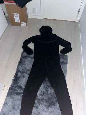 Svart Nike tracksuit  - Säljer en Nike svart tracksuit med huva och hel dragkedja framtill. Den har långa ärmar, smal passform och reflexdetaljer över bröstet och på ena ärmen. Perfekt för chill dagar eller när du vill vara bekväm men ändå se cool ut.