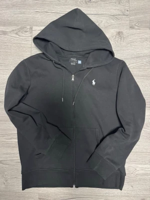 Svart hoodie från Polo Ralph Lauren - Snygg svart hoodie med dragkedja från Polo Ralph Lauren. Klassisk design med huva och dragsnören, samt den ikoniska vita logon broderad på bröstet. Tillverkad i mjuk bomullsblandning och har två fickor framtill. Perfekt för en avslappnad streetwear-look.