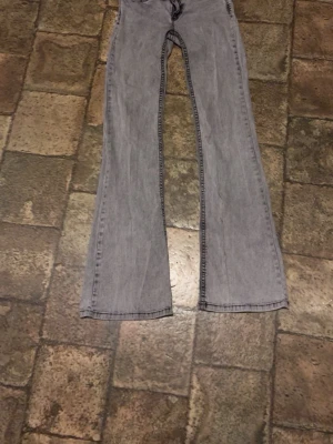 Ljusgrå bootcut jeans med broderi - Säljer ett par ljusgrå bootcut jeans med snyggt broderade bakfickor. Jeansen har en klassisk femficksmodell och är tillverkade i denim med lätt tvättad look. Perfekta för dig som gillar retrostil och vill ha ett par jeans med unik detalj på fickorna.