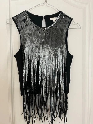 Svart paljettopp med fransar - Supersnygg svart ärmlös topp med silverfärgade paljetter och långa fransar framtill och baktill. Toppen har rund halsringning och knäppning i nacken. Perfekt för dig som vill sticka ut med extra bling och rörelse i outfiten.