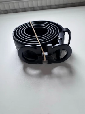 Svart skärp från Salvatore Ferragamo - Snyggt svart skärp från Salvatore Ferragamo med ikoniskt dubbel-Gancio-spänne i matt svart metall. Bältet är gjort i präglat skinn och har en stilren, modern look som passar till många outfits. Perfekt accessoar för dig som gillar exklusiva detaljer.