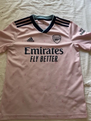 Arsenal rosa matchtröja  - Säljer en rosa Arsenal matchtröja från Adidas med mörkblå detaljer och logga på bröstet.  Tröjan är tredje tröjan 2022/23. Tröjan har korta ärmar, V-ringning och små diskreta mönster över hela tyget. Perfekt för fotboll eller träning. Har en liten fläck men ger inget tydligt.