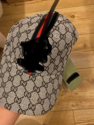 Grå keps med GG-mönster och ränder - Snygg grå keps med all-over GG-mönster i svart och klassiska gröna och röda ränder framtill. Kepsen har böjd skärm och justerbar rem bak. Materialet känns slitstarkt och har en exklusiv känsla, perfekt för dig som gillar streetwear och designerinspirerat.