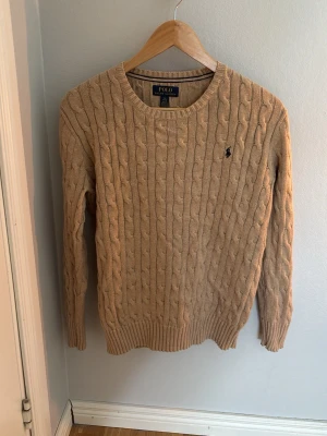 Beige kabelstickad tröja Polo Ralph Lauren - Kabelstickad beige tröja från Polo Ralph Lauren med rund halsringning och den klassiska broderade loggan på bröstet. Tröjan har långa ärmar och ribbade muddar vid ärmslut och nederkant. Perfekt för lager på lager och en clean look.