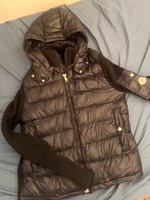 Svart dunväst med huva från moncler - Snygg svart dunväst med avtagbar huva och stickade ärmar från moncler. Västen har dragkedja framtill, två fickor med tryckknappar och ribbstickade detaljer vid ärmslut och krage. Perfekt för lager-på-lager och chill stil.