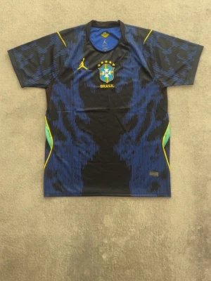 Blå Brasilien matchtröja - Snygg blå och svart matchtröja med Brasilien-emblem på bröstet och gula detaljer. Tröjan har korta ärmar, rund hals och inslag av grönt och gult på sidorna. Tillverkad i ett lätt och ventilerande material, perfekt för fotboll eller träning.