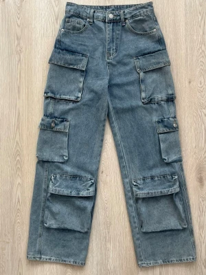 Baggy cargobyxor i denim - Säljer ett par baggy cargobyxor i ljusblå denim med flera stora fickor på benen och klassisk cargomodell. Byxorna har raka ben, bälteshällor och knappgylf. Perfekta för en avslappnad streetwear-look.