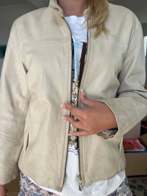 Beige skinnjacka med dragkedja - Snygg beige skinnjacka med rak passform och hög krage. Jackan har diskreta sömdetaljer över bröstet och längs ärmarna samt en stilren dragkedja framtill. Perfekt för dig som vill ha en clean och tidlös look.