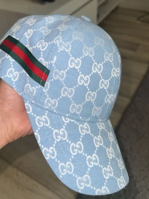 Ljusblå Gucci keps med GG-mönster - Snygg ljusblå keps från Gucci med klassiskt vitt GG-mönster över hela kepsen. På sidan finns en grön och röd rand som signaturdetalj. Justerbar rem baktill med metallspänne. Tillverkad i tygmaterial som känns både trendigt och exklusivt.
