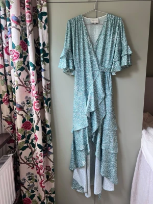 Blommig långklänning från By Malina - Ljusblå långklänning med små vita blommor från By Malina. Klänningen har omlottmodell, volangdetaljer och v-ringning. Ärmarna är korta med dubbla volanger och klänningen har en flowy passform, perfekt för sommarens alla tillfällen.