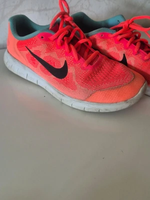 Nike rosa sneakers med svart swoosh - Snygga sneakers från Nike i en stark rosa färg med svart swoosh på sidan. Ovandelen är i mesh för bra andningsförmåga och skorna har vita sulor. Insidan är turkosblå och skorna har rosa snören. Perfekta för dig som vill sticka ut med färg på fötterna.