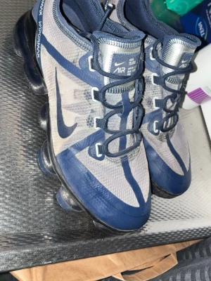Nike Air Vapormax blå sneakers - Nike Air Vapormax sneakers i blått och grått med genomskinlig mesh-överdel och tydliga blå detaljer. Skorna har en unik sula med synliga luftbubblor och klassisk Nike Swoosh på sidan. Låga och sportiga med snörning och modern design.