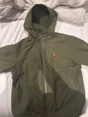Olivgrön Ralph lauren vindjacka med huva  - Snygg olivgrön vindjacka med huva och dragkedja framtill. Jackan har en liten röd logga på bröstet och justerbar dragsko i huvan. Perfekt för blåsiga dagar och enkel att matcha med olika outfits.