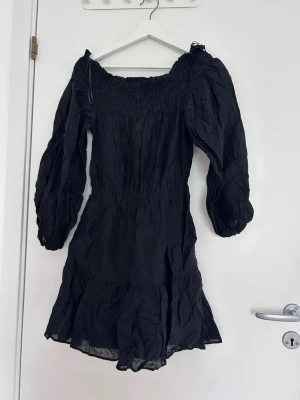 Svart offshoulder klänning - Trendig svart kortklänning med offshoulder-design och långa puffärmar. Klänningen har smock upptill och volang nedtill, vilket ger en snygg och feminin siluett. Perfekt för dig som gillar en stilren look med lite edge.