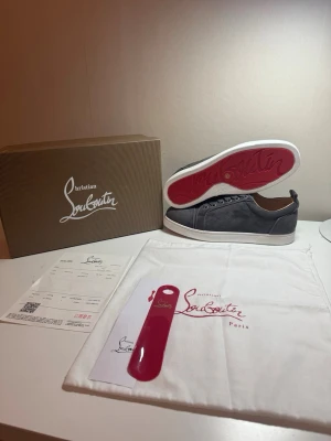 Gråa Christian Louboutin sneakers - Snygga gråa sneakers från Christian Louboutin med klassisk röd sula och vita kanter. Skorna har lågt skaft, snörning och är tillverkade i mocka med beige innersida. Perfekta för dig som vill sticka ut med exklusiv stil.