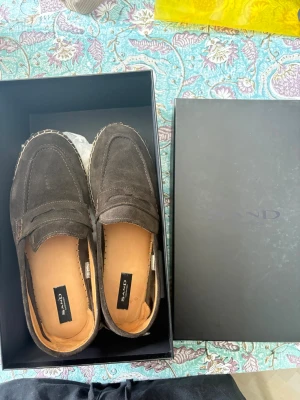 Bruna mocka loafers från SAND - Snygga bruna loafers i mocka från SAND. Klassisk penny loafer-design med rundad tå och dekorativ söm runt ovandelen. Skorna har en ljus innersula och är perfekta för en stilren och avslappnad look.
