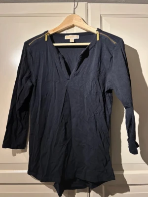 Svart blus med dragkedjedetaljer Micheal Kors - Stilren svart blus från Michael Kors med långa ärmar och snygga guldfärgade dragkedjor på axlarna. Blusen har en djup V-ringning och är tillverkad i ett mjukt tyg som ger en avslappnad look. Perfekt att matcha med jeans eller kjol. storlek M.