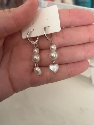 Silvriga örhängen med pärlor och hjärta - Snygga örhängen i silverfärg med hängande vita pärlor och ett hjärtformat hänge längst ner. Perfekt accessoar för att lyfta din stil med en söt och trendig touch. Passar dig som gillar unika detaljer och pärlor.