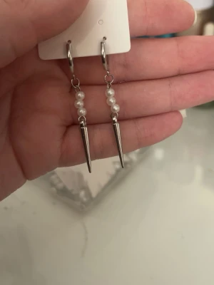 Silvriga örhängen med pärlor och spikes - Säljer ett par coola örhängen i silverfärg med små vita pärlor och spetsiga spikes. Örhängena har en modern och edgy design som sticker ut. Perfekta för dig som gillar att mixa sött och tufft i din stil.
