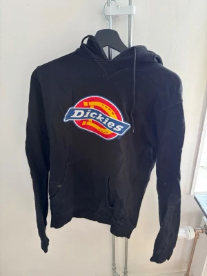 Svart Dickies hoodie med logga - Säljer en svart hoodie från Dickies i storlek XS. Hoodien har en stor färgglad Dickies-logga på bröstet, huva med dragsko och en klassisk känguruficka framtill. Perfekt för dig som gillar streetwear och vill ha en enkel men snygg look.