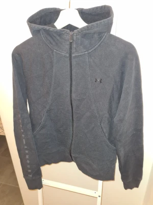 Svart hoodie från Under Armour - Snygg svart hoodie från Under Armour med dragkedja framtill och huva. Diskret logga på bröstet och text längs ena ärmen. Mjuk och bekväm med ribbade detaljer vid fickorna. Perfekt för en sportig och avslappnad stil. Liten i storlek.