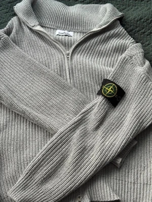 Ljusgrå stickad tröja från Stone Island - Snygg ljusgrå stickad tröja från Stone Island med dragkedja framtill och hög krage. Tröjan har ribbstickade muddar och den ikoniska Stone Island-loggan på ärmen. Perfekt för dig som gillar streetwear och vill ha något både stilrent och bekvämt.