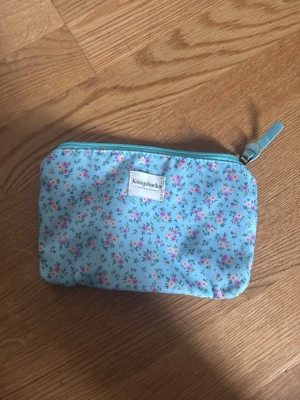 Blommig necessär från Kungshamn - Söt liten necessär i ljusblått tyg med blommigt mönster i rosa, grönt och vitt. Dragkedja upptill med matchande blått band. Perfekt för småsaker eller smink. Märkt med Kungshamn på framsidan.