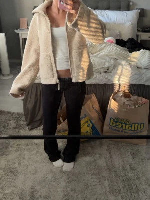 Beige teddyjacka med dragkedja - Mysig beige teddyjacka med dragkedja framtill och två stora fickor. Jackan har en hög krage och är gjord i fluffigt, mjukt material som ger en avslappnad och trendig look. Perfekt för kyliga dagar och enkel att matcha med olika outfits.