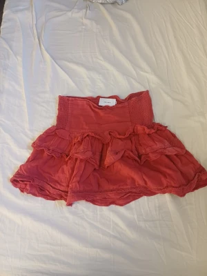 Röd volangkjol med smockdetalj - Supersöt kort kjol i rosa med bred smock i midjan och flera lager volanger. Kjolen har en luftig och flowy känsla, perfekt för varma dagar. Enkel att matcha med en crop top eller t-shirt. Skulle säga S-M i passformen