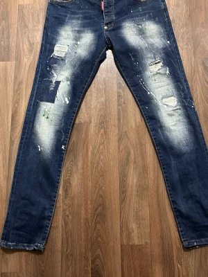 Mörkblå DSQUAREDS2 - Säljer ett par mörkblå jeans med slitna detaljer, patchar och färgstänk i vitt och grönt. Jeansen har raka ben, klassisk femficksmodell och tydliga slitningar både fram och bak. Perfekt för dig som gillar en edgy och kreativ stil.