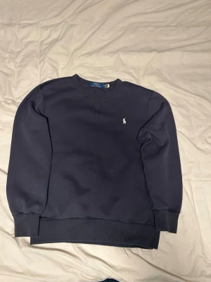 Mörkblå sweatshirt från Polo Ralph Lauren - Klassisk mörkblå sweatshirt från Polo Ralph Lauren med rund halsringning och broderad vit logga på bröstet. Tröjan har ribbade muddar vid ärmslut och nederkant. Perfekt för en avslappnad och stilren look.