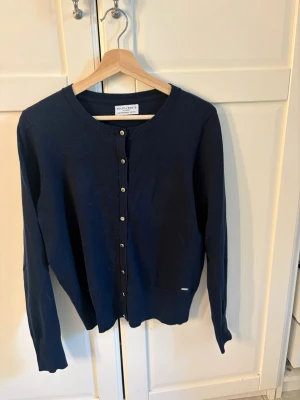 Mörkblå kofta från Holly & Whyte - Klassisk mörkblå kofta från Holly & Whyte by Lindex med rund halsringning och knappar framtill. Tillverkad i mjukt stickat material och har långa ärmar samt ribbade muddar vid ärmslut och nederkant. Perfekt att slänga över en t-shirt eller blus.