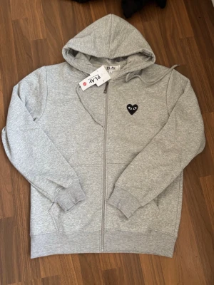 Comme Des Garçons - Play Grey hoodie zip - Snygg grå zip-hoodie med huva och dragsko. På bröstet finns ett litet svart hjärta med ögon och på ryggen ett större hjärta i samma stil. Mjuk bomullsblandning och ribbade muddar. Perfekt för en chill och trendig look.