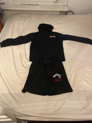 Svart Hoodrich hoodie och shorts set - Säljer ett svart set från Hoodrich med hoodie och matchande shorts. Hoodien har tryck i vitt och rött på bröstet och ryggen, samt huva. Shortsen har dragsko i midjan och tryck på ena benet. Perfekt för en avslappnad streetwear-look.