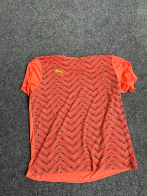 Neonorange träningströja från Puma - Snygg neonorange träningströja från Puma med korta ärmar och svart zigzag-mönster på framsidan. Tillverkad i ett lätt och snabbtorkande material som passar perfekt till träning eller löpning. Puma-logga i gult på bröstet.