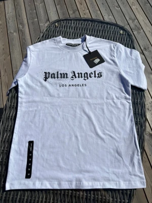 Vit Palm Angels t-shirt Los Angeles - Snygg vit t-shirt från Palm Angels med svart tryck 'Palm Angels Los Angeles' på bröstet. Klassisk rund hals och korta ärmar. Perfekt för dig som gillar streetwear och vill ha en clean look med tydlig logga.