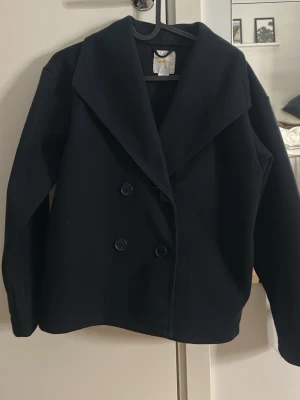 Marinblå skepparkavaj med bred krage - Stilren marinblå skepparkavaj med dubbelknäppning och bred krage. Klassisk rak passform och två sidofickor. Perfekt för dig som gillar tidlös och enkel stil. Materialet ser ut att vara ullblandning för extra värme. Passar dig som e storlek xs 