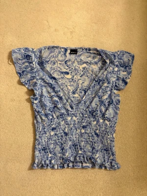 Blåvit blus med volang och v-ringning - Supersöt blåvit blus med v-ringning och volangdetaljer vid ärmar och nederkant. Blusen har ett blommigt mönster och är lätt rynkad för en smickrande passform. Perfekt till jeans eller kjol för en fräsch look.