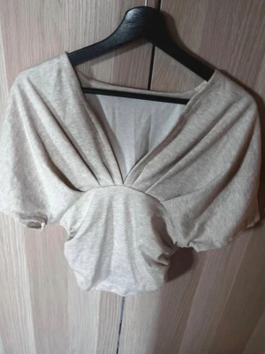 Beige croppad topp med v-ringning - Trendig beige croppad topp med djup v-ringning och korta ärmar. Tillverkad i mjukt bomullsmaterial och har en snygg draperad effekt framtill. Perfekt för att styla med höga jeans eller kjol.