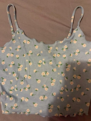 Blommig ljusblå ribbad linnetopp - Supersöt ljusblå linnetopp med små vita blommor och gröna detaljer. Toppen är ribbad med smala axelband och vågig kant upptill och nedtill. Perfekt för en somrig look och enkel att matcha med jeans eller kjol.