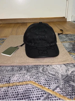 Svart keps från Gucsi med GG-mönster - Snygg svart keps från Gucsi med det klassiska GG-mönstret över hela kepsen. Tillverkad i mesh-liknande material med böjd skärm och justerbar rem bak. Perfekt accessoar för en trendig och exklusiv look.