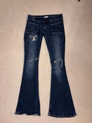 Mörkblå bootcut jeans med slitningar - Säljer ett par mörkblå bootcut jeans med tydliga slitningar och hål på låren och knäna. Jeansen har låg midja, fem fickor och en klassisk dragkedja. Perfekta för en avslappnad och trendig look.