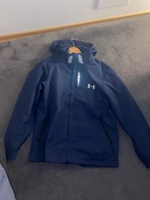 Mörkblå vindjacka från Under Armour - Snygg mörkblå vindjacka från Under Armour med huva och dragkedja framtill. Jackan har två snedställda fickor med dragkedja och reflekterande detaljer på kragen. Tillverkad i ett lätt och vattenavvisande material, perfekt för blåsiga dagar.