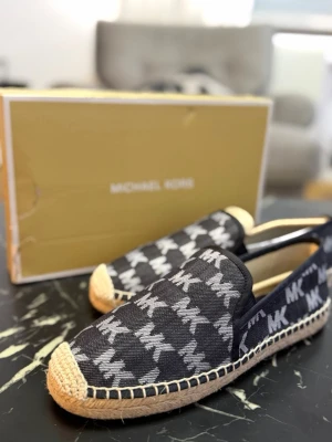 Svarta loafers från Michael Kors - Snygga svarta loafers från Michael Kors med vit logotypprint över hela skon. Ovandelen är i textil och sulan har en flätad detalj i naturfärgad jute. Skorna har en klassisk slip-on design och är perfekta för en trendig och avslappnad look.