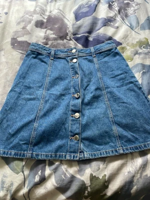 Blå jeanskjol med knappar framtill - Säljer en klassisk blå jeanskjol i kort modell med silverfärgade knappar längs hela framsidan. Kjolen har hällor för skärp och en lätt utställd passform. Perfekt till vardags för en avslappnad och trendig look.