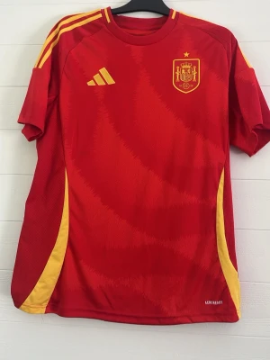 Spaniens röd fotbollströja Adidas - Röd matchtröja från Adidas för Spaniens landslag med gula detaljer och emblem på bröstet. Tröjan har korta ärmar, rund hals och gula ränder på axlarna. Tillverkad i lätt och ventilerande material, perfekt för träning eller match.